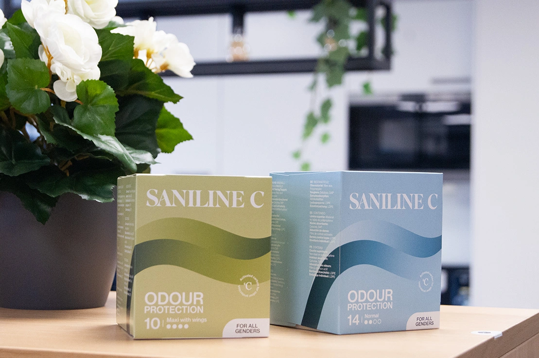 Två förpackningar av inkontinensskydd Saniline C Odour Protection står bredvid en krukväxt. En av förpackningarna är grön och den andra blå och innehåller aktivt kol som har en lukthämmande effekt.