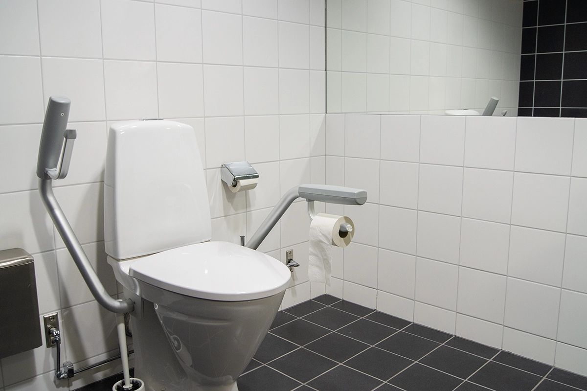 Vit toalettstol med grå stödhandtag i ett badrum med vitt och svart kakel.