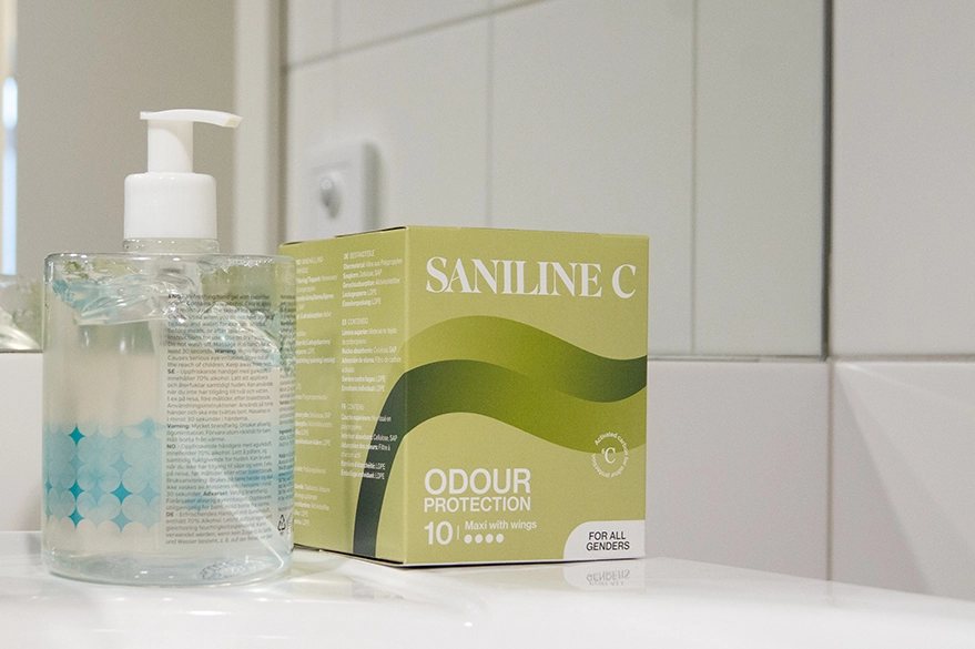 Grön förpackning med Saniline C Maxi with wings och en tvål flaska i ett badrum. Ett bra hjälpmedel mot lukt och läckage vid sfinkterskada.