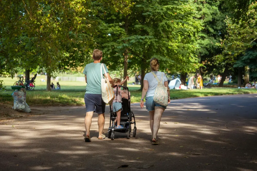 En familj som går ute i en park. Det är en mamma, pappa och ett barn. Barnet står på en barnvagn.