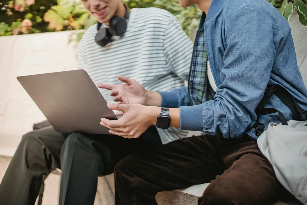 Två men sitter ute på en bänk och pratar framför en laptop.